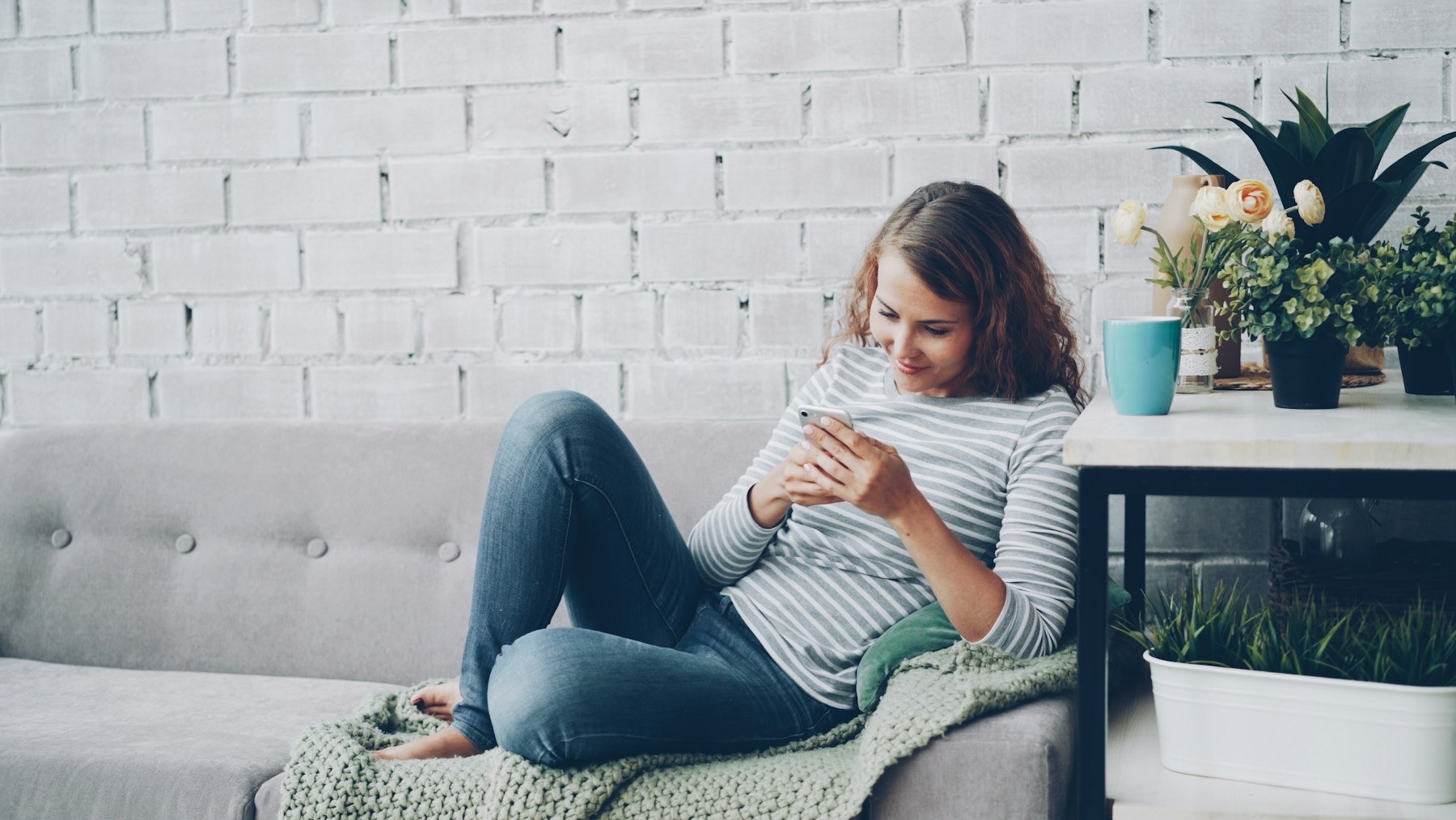 A woman relaxes on a grey sofa at home, smiling at her phone, with plants and a mug on the side table.