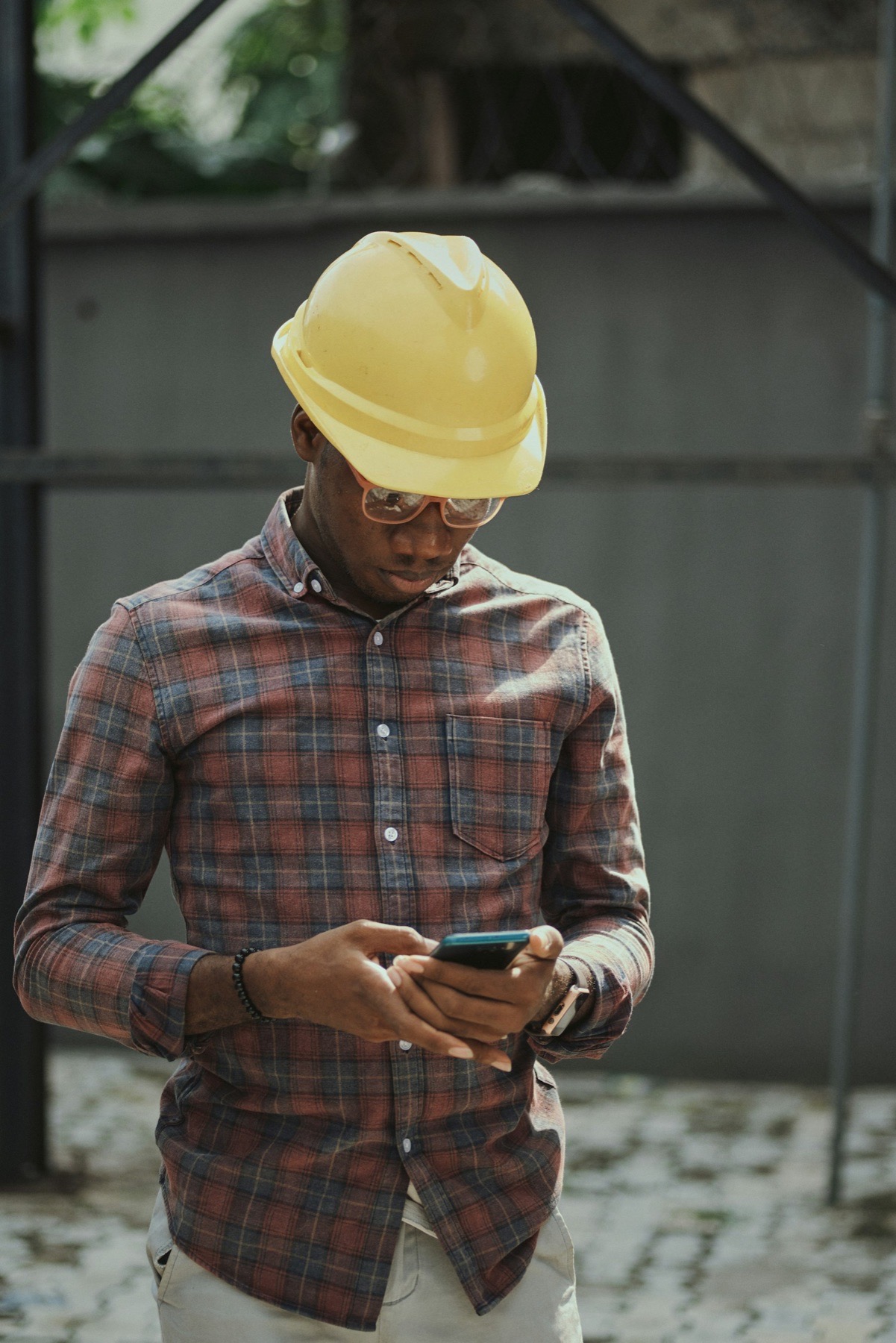 A builder in a yellow hard hat and checked shirt stands on site in daylight, looking down at his phone.