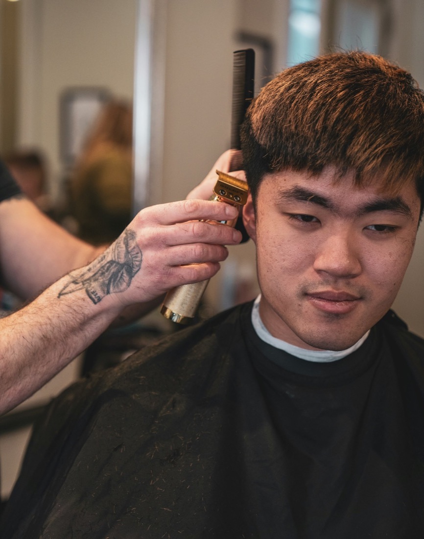 A barber trimming a client's hair with clippers in a bright barbershop.