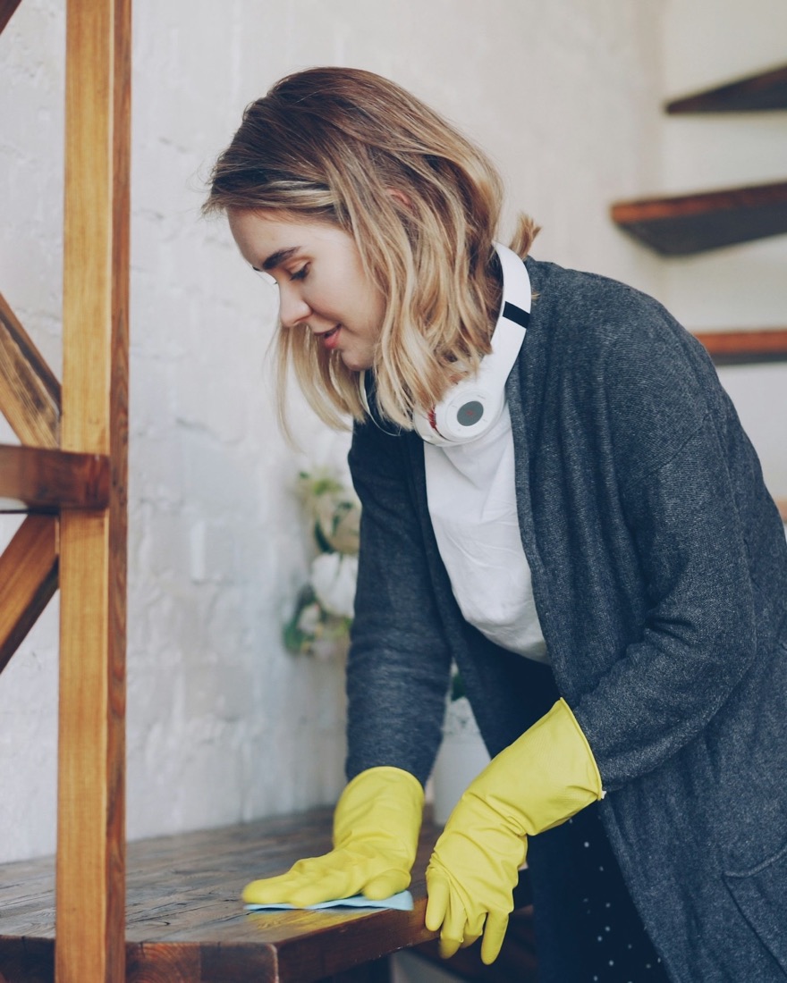 A cleaner in yellow gloves wiping a wooden shelf in a bright home.