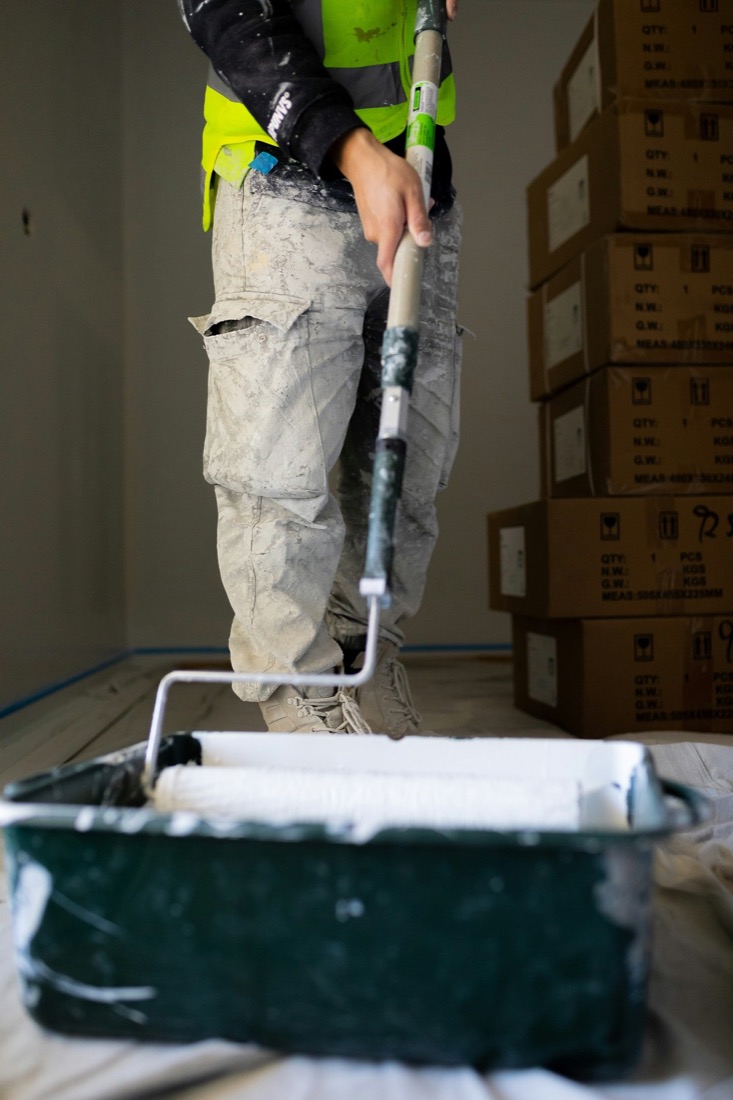 A decorator in paint-spattered overalls loading a roller from a tray on a dust sheet.
