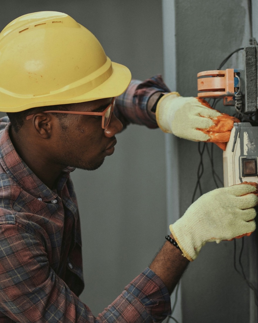 An electrician in a yellow hard hat and gloves fitting a socket on a grey wall.
