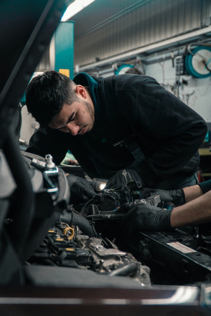 A mechanic leaning under an open bonnet with a torch, working on the engine.